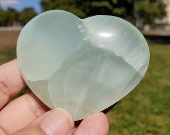 May include: A pale green heart-shaped stone, held in a hand, against a backdrop of green grass and a blue sky. The stone has a smooth, polished surface with subtle variations in colour and texture, suggesting it is a natural gemstone.