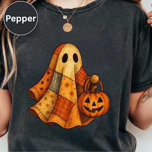May include: A charcoal gray t-shirt featuring a graphic of a ghost draped in a patchwork quilt made of orange, yellow, and brown fabric squares with star and floral patterns. The ghost is holding a small orange jack-o'-lantern bucket.