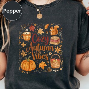 May include: A charcoal gray t-shirt featuring a graphic print with the text "Cozy Autumn Vibes" surrounded by illustrations of a pumpkin, a mug, a squirrel, a basket of apples, stacked blankets, and various autumn leaves in orange and brown.