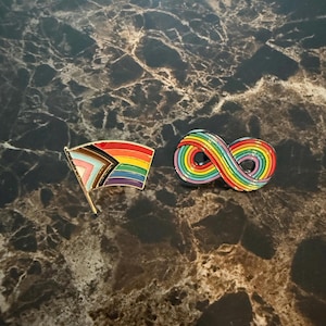 Puede incluir: Dos pines de esmalte sobre una superficie de mármol. Un pin es una bandera arcoíris con un asta dorada. El otro pin es un símbolo de infinito arcoíris. Ambos pines son de los colores de la bandera del orgullo.