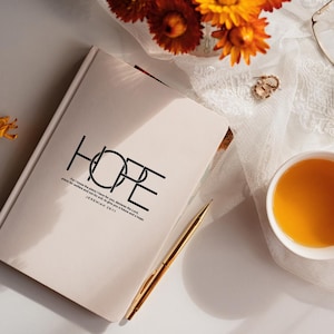 Op de afbeelding: Een crèmekleurig notitieboek met het woord "HOPE" in zwart gedrukt, samen met een bijbelvers. Een gouden pen ligt naast het notitieboek. De afbeelding bevat ook een kopje thee, oorbellen en bloemen.