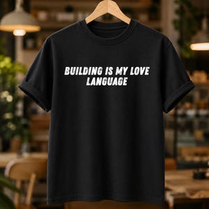 Peut inclure: Un t-shirt noir à manches courtes avec le texte blanc au style vieilli "BUILDING IS MY LOVE LANGUAGE" imprimé sur la poitrine. Le vêtement possède un col rond et est présenté sur un cintre en bois devant un arrière-plan flou.