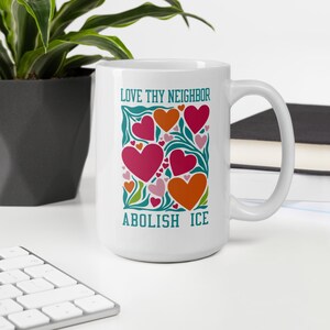 Peut inclure: Mug en céramique blanche avec le texte "LOVE THY NEIGHBOR" et "ABOLISH ICE" en turquoise. Le mug présente un motif de cœurs roses et oranges, entourés de feuilles et de vignes vertes.
