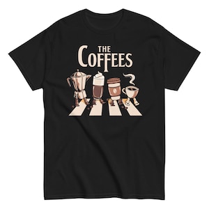 Peut inclure: T-shirt noir avec un graphisme représentant des personnages sur le thème du café traversant une rue, avec le texte "The Coffees" au-dessus. Les personnages incluent une cafetière, un latte, un gobelet de café à emporter et une tasse à café.