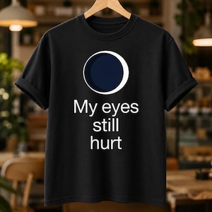 Peut inclure: Un t-shirt noir à manches courtes orné d'un graphique représentant une phase de lune croissante au-dessus du texte blanc "My eyes still hurt". Le t-shirt possède un col rond et est présenté sur un cintre en bois, affichant un design simple.