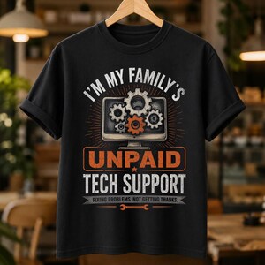 Op de afbeelding: Een zwart t-shirt met korte mouwen en een afbeelding van een computermonitor met tandwielen. De tekst luidt: I'M MY FAMILY'S UNPAID TECH SUPPORT. FIXING PROBLEMS. NOT GETTING THANKS. Het ontwerp bevat een oranje tandwiel en een moersleutel.