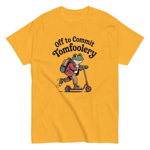 Può includere: T-shirt giallo senape con un disegno di una rana su un monopattino rosso. La rana indossa uno zaino e una giacca rossa. Il testo sopra la rana dice "Off to Commit Tomfoolery". Un design divertente e giocoso.