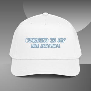 May include: A white baseball cap featuring the text "BUILDING IS MY LOVE LANGUAGE" printed in a light blue, stylized font across the front panel. The cap has a structured crown, a curved brim, and a button detail at the top center.