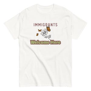 Może przedstawiać: Biała koszulka z napisem "IMMIGRANTS Welcome Here" w kolorze brązowym i zielonym. Projekt zawiera kwiat i trzy motyle w odcieniach pomarańczu i brązu.