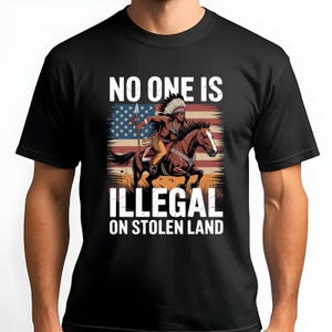 Może przedstawiać: Czarna koszulka z wizerunkiem Indianina na koniu na tle amerykańskiej flagi. Na koszulce widnieje biały napis "NO ONE IS ILLEGAL ON STOLEN LAND".