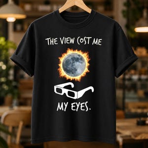 Peut inclure: Un t-shirt noir à manches courtes avec un motif représentant une lune entourée de flammes orange au-dessus d'une paire de lunettes d'éclipse blanches. Le texte "THE VIEW COST ME" est imprimé au-dessus et "MY EYES." en dessous en blanc.