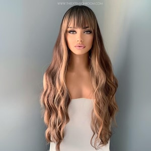 May include: A long, wavy wig with a dark brown base and blonde highlights. The wig has a blunt fringe and is styled with loose curls. The image includes the website address: www.theextensionroom.com.