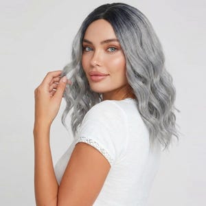 May include: A shoulder-length wig featuring wavy, silver-gray hair with dark roots. The hair has a center part and a soft, layered texture that frames the face. The wig is styled with loose, natural-looking waves throughout the length.