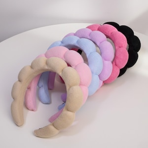 Pode incluir: Uma coleção de tiaras acolchoadas em várias cores pastel, incluindo bege, rosa, azul claro, lilás, rosa choque e preto. Cada tiara apresenta uma série de segmentos arredondados e acolchoados, criando uma aparência texturizada e semelhante a uma nuvem. As tiaras estão dispostas em fila.