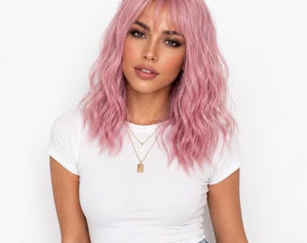 Puede incluir: Una peluca ondulada a la altura de los hombros en un tono rosa pastel suave con flequillo recto que enmarca la frente. El cabello tiene una textura en capas con ondas sueltas, creando un aspecto voluminoso que cae más allá de los hombros.