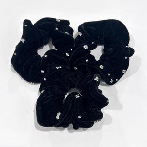 May include: Four black velvet scrunchies with small, sparkling, clear rhinestones scattered across the surface. The scrunchies are arranged in a circular pattern against a white background. These hair accessories add a touch of elegance.