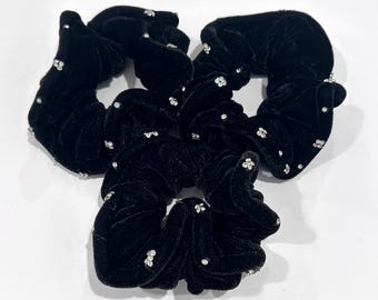 May include: Four black velvet scrunchies, each adorned with tiny, clear rhinestones. The scrunchies are arranged in a circular pattern against a white backdrop. These hair accessories are a stylish addition to any hairstyle.