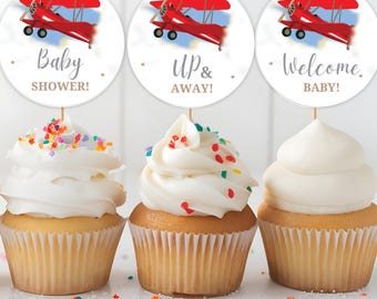 May include: Three cupcakes with white frosting and colorful sprinkles. Each cupcake has a circular topper with a red biplane design and text that reads "Baby Shower!", "Up & Away!", and "Welcome, BABY!"