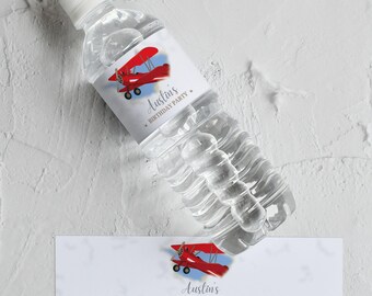 May include: Clear plastic water bottle with a white cap and a custom label. The label features a red biplane illustration with the text "Austin's BIRTHDAY PARTY". A matching label is below the bottle. The bottle is on a white textured surface.