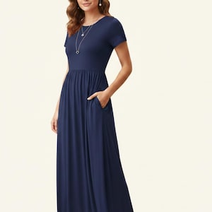 May include: A navy blue maxi dress with short sleeves and a round neckline. The dress has a gathered waist and side pockets. A silver necklace is worn. The model is wearing pearl earrings.
