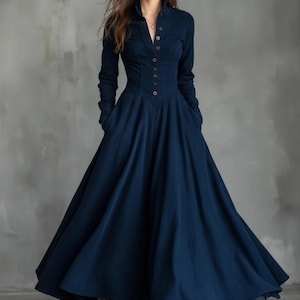 Può includere: Un lungo abito blu navy con collo alto e maniche lunghe. L'abito presenta una chiusura a bottoni sul davanti e una gonna svasata. L'orlo è rifinito con un delicato dettaglio in pizzo. L'abito ha tasche.