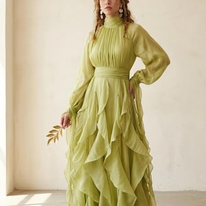 May include: A long, flowing, light green dress with a high neckline and long sleeves. The dress features a layered, ruffled skirt and a matching belt. The model is holding a gold leaf. The dress is made of a lightweight, sheer fabric.