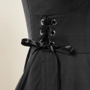 May include: Close-up of a black garment featuring a corset-style back with silver eyelets and a black satin ribbon. The garment has a fitted bodice and a flared skirt, suggesting a dress or top.