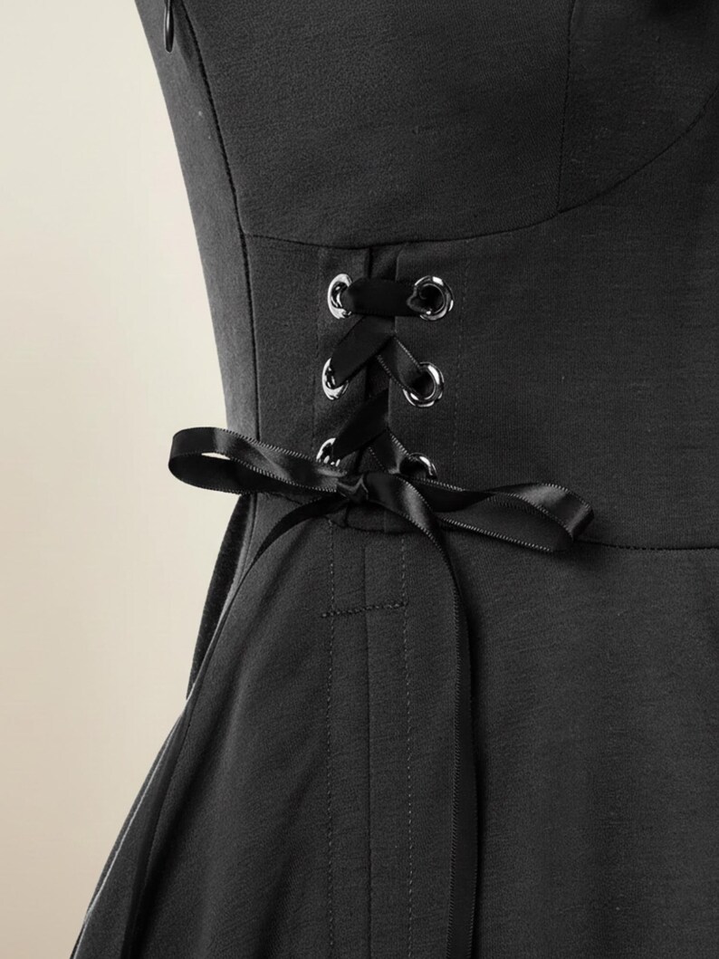 May include: Close-up of a black garment featuring a corset-style back with silver eyelets and a black satin ribbon. The garment has a fitted bodice and a flared skirt, suggesting a dress or top.