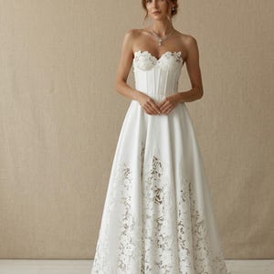 May include: A strapless white wedding dress with a fitted bodice and a full skirt. The bodice features floral lace detailing, and the skirt has a lace overlay at the hem. The model is wearing a necklace.