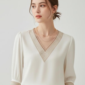 Könnte beinhalten: Eine cremefarbene Bluse mit V-Ausschnitt und Dreiviertelärmeln. Der Ausschnitt und die Ärmelbündchen weisen ein detailliertes, gesticktes Design auf. Die Bluse wird mit einer beigefarbenen Hose und goldfarbenem Schmuck kombiniert.