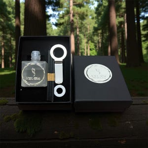May include: A black gift box set containing a frosted glass bottle with a logo, a silver bottle opener, and black reeds. The box is open, revealing the contents, set against a forest backdrop. The bottle is square.