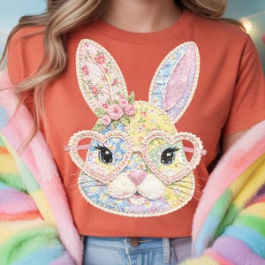 May include: Orange t-shirt featuring a patchwork bunny face with heart-shaped glasses. The bunny has floral ears and a nose, with a cluster of pink roses. The shirt is paired with blue jeans and a colorful, rainbow-striped jacket.