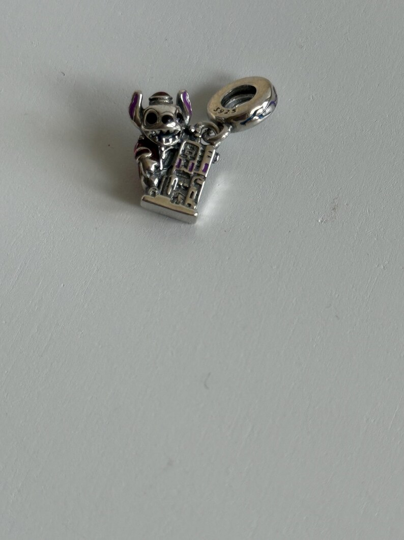 Puede incluir: Un charm de plata con el personaje Stitch de la pel&iacute;cula Lilo & Stitch. El charm muestra a Stitch sosteniendo un edificio, con detalles morados en sus orejas y ropa. El charm tiene un lazo para sujetarlo a una pulsera o collar.