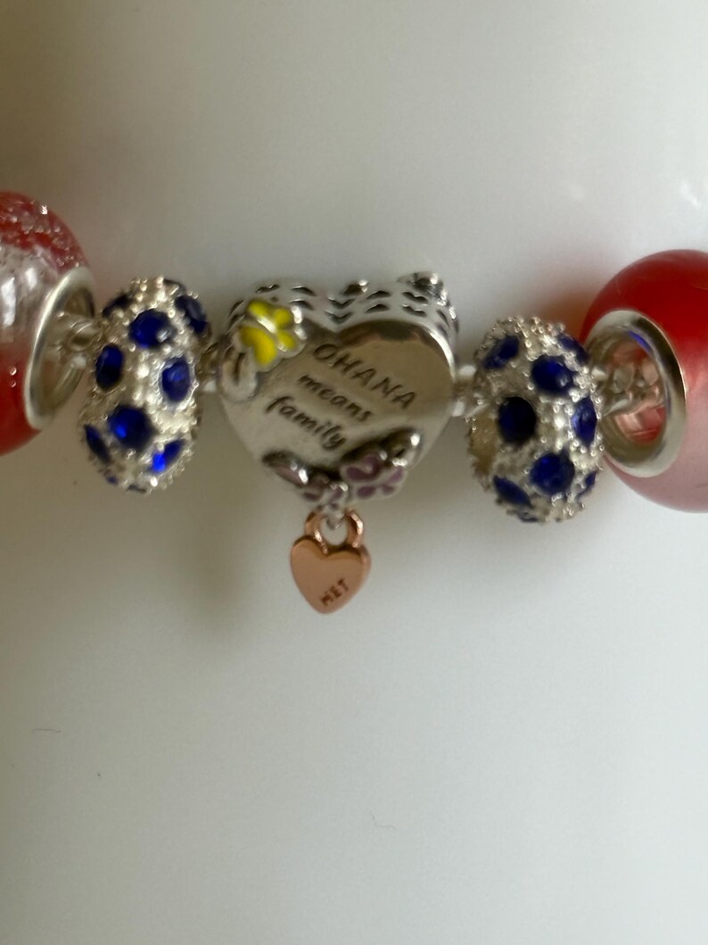 Puede incluir: Una pulsera de abalorios con un dije en forma de coraz&oacute;n que dice "OHANA means family". La pulsera tiene cuentas rojas y transparentes, cuentas plateadas con gemas azules y un peque&ntilde;o dije de coraz&oacute;n de oro rosa.