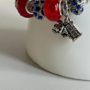 Puede incluir: Una pulsera de plata con abalorios rojos y azules. La pulsera presenta un dije de un personaje de dibujos animados y varias cuentas con piedras preciosas azules. La pulsera est&aacute; sobre una superficie blanca.