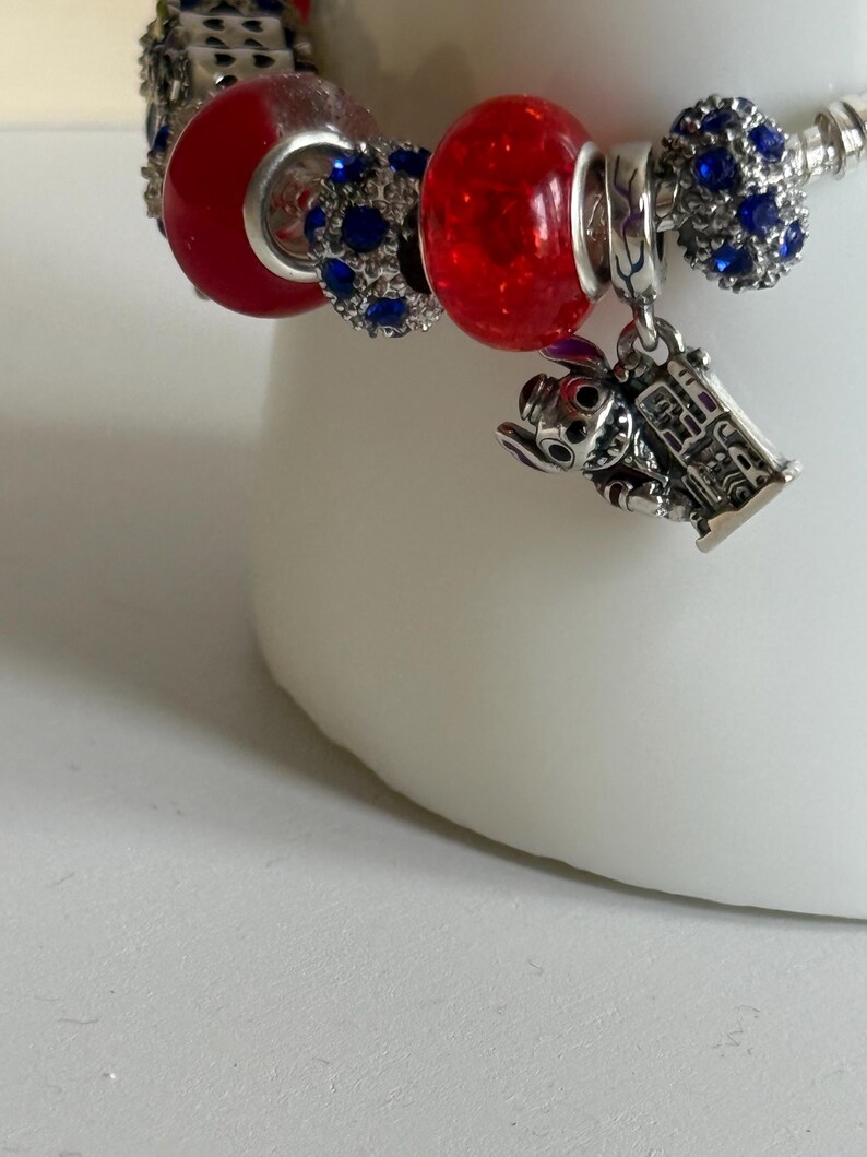 Puede incluir: Una pulsera de plata con abalorios rojos y azules. La pulsera presenta un dije de un personaje de dibujos animados y varias cuentas con piedras preciosas azules. La pulsera est&aacute; sobre una superficie blanca.
