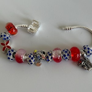 Puede incluir: Una pulsera de plata con varios abalorios decorativos. Los abalorios incluyen cuentas rojas y azules, una flor roja y un abalorio con un dise&ntilde;o de castillo. Otros abalorios tienen un dise&ntilde;o de Mickey Mouse.