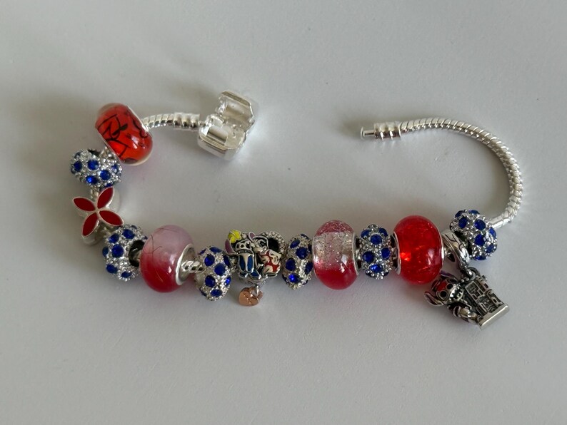 Puede incluir: Una pulsera de plata con varios abalorios decorativos. Los abalorios incluyen cuentas rojas y azules, una flor roja y un abalorio con un dise&ntilde;o de castillo. Otros abalorios tienen un dise&ntilde;o de Mickey Mouse.