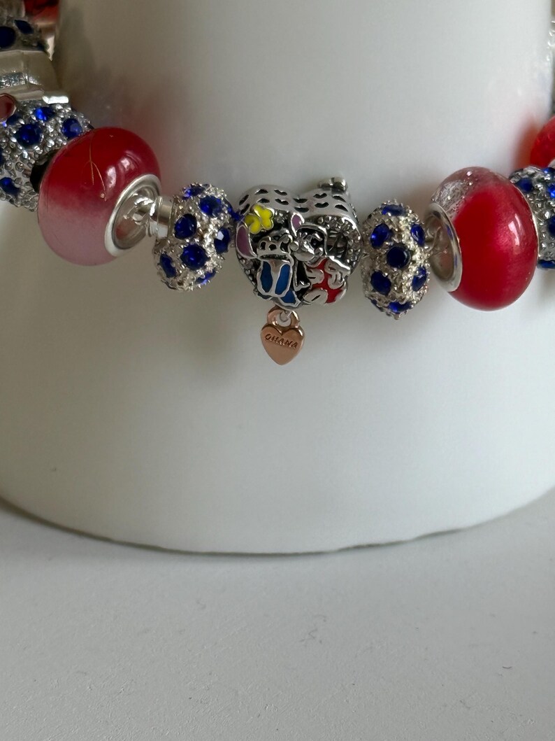 Puede incluir: Una pulsera de abalorios con varias cuentas. La pulsera incluye cuentas rojas y azules, junto con un dije que representa un personaje de dibujos animados. Tambi&eacute;n hay un dije en forma de coraz&oacute;n con la palabra "Ohana".