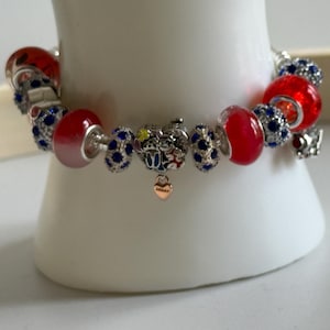 Puede incluir: Una pulsera de plata con abalorios rojos y azules. La pulsera presenta cuentas de vidrio rojas, cuentas azules con incrustaciones de cristal y un peque&ntilde;o dije de coraz&oacute;n dorado. La pulsera se exhibe sobre una superficie blanca.