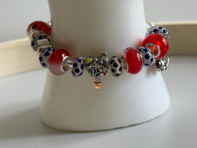 Puede incluir: Una pulsera de abalorios plateada con cuentas rojas y azules. La pulsera presenta cuentas de vidrio rojas, cuentas azules con incrustaciones de cristal y un peque&ntilde;o dije en forma de coraz&oacute;n. La pulsera se exhibe sobre una superficie blanca.