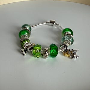 May include: A silver charm bracelet with various green and silver beads. The bracelet features several charms, including a four-leaf clover and a small horse. The beads are different shades of green, with some having a textured appearance.