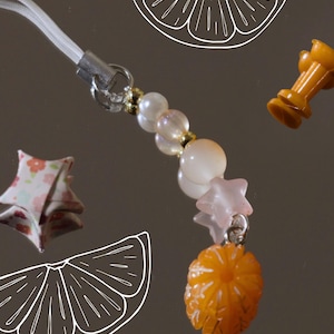 May include: A white string phone charm with a silver clasp, beads, and a pink star charm. The charm also features a small orange citrus fruit charm.