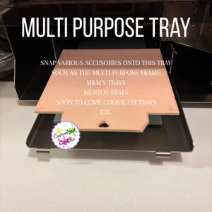 以下が含まれることがあります： 「MULTI PURPOSE TRAY」の文字が入った多目的トレイ。トレイは薄いピンク色で、濃い金属製のベースの上に置かれています。画像には、M&M'sやMentosのトレイなど、アクセサリーをトレイにスナップできると書かれています。「Customised Cookies」のロゴが隅にあります。