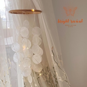May include: A decorative hanging mobile with a wooden ring, strings of small, round, white shell-like discs, and tiny fairy lights. The mobile hangs in front of a sheer curtain with embroidered floral patterns. The text "bright revival HOME DECOR GIFTS" is visible.