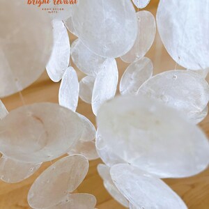 May include: A close-up of a decorative mobile featuring numerous, translucent, oval-shaped, white shell discs. The discs are strung together with thin, clear string. The mobile is suspended, creating a layered effect. The text "bright revival HOME DECOR GIFTS" is visible.