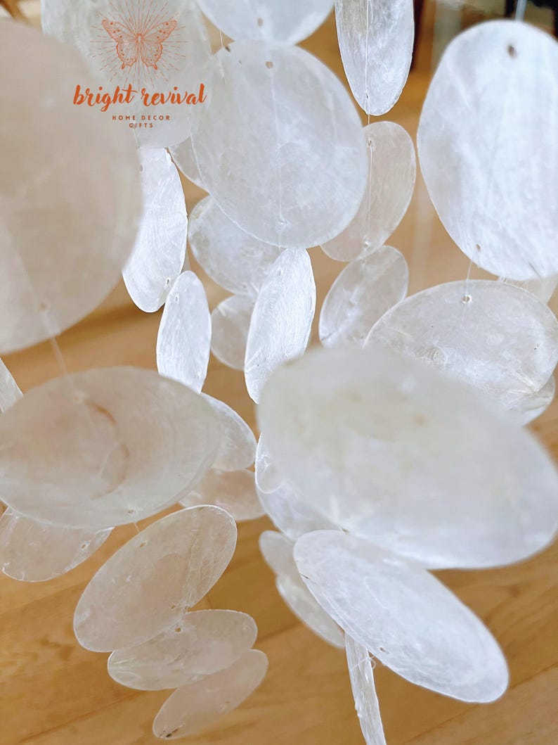 May include: A close-up of a decorative mobile featuring numerous, translucent, oval-shaped, white shell discs. The discs are strung together with thin, clear string. The mobile is suspended, creating a layered effect. The text "bright revival HOME DECOR GIFTS" is visible.