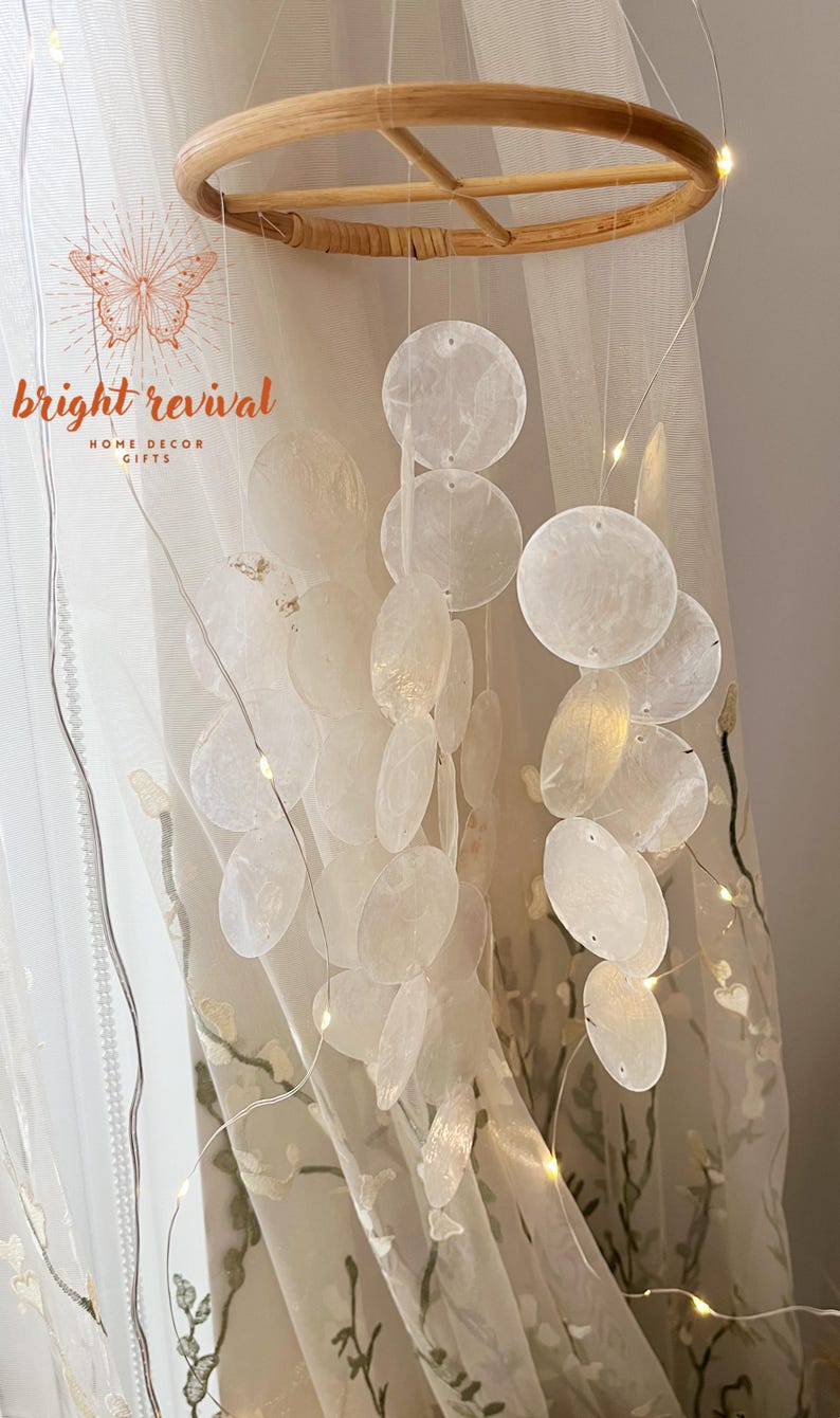 May include: A decorative mobile featuring a natural wood ring and numerous white, disc-shaped elements suspended by thin strings. The mobile hangs in front of a sheer curtain, with the text "bright revival HOME DECOR GIFTS" visible.