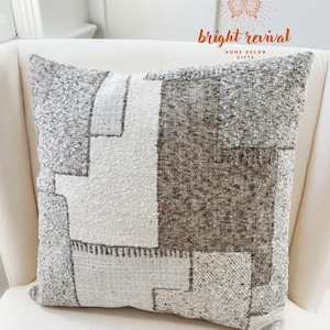 May include: Square cushion with a geometric patchwork design in shades of grey, brown, and cream. The cushion is made of textured fabric and is labelled "bright revival HOME DECOR GIFTS".
