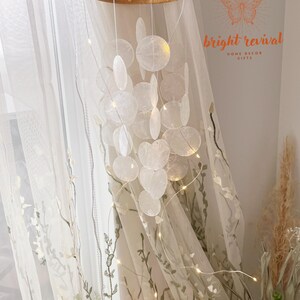 May include: A decorative wind chime featuring iridescent, shell-shaped pendants suspended from a wooden ring. The chime is illuminated by small, warm white fairy lights. The background includes sheer curtains and the text "bright revival HOME DECOR GIFTS".